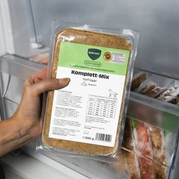 Hand hält verpacktes tiefgefrorenes Hundefutter mit Aufschrift 'Komplett‑Mix Geflügel' (BARFER'S Komplett), 500 g.