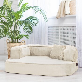 Ein plüschiges, cremefarbenes Haustierbett mit einer unterstützenden Rückenlehne und einem kleinen Kissen, in einem hellen Raum mit einer Topfpflanze.