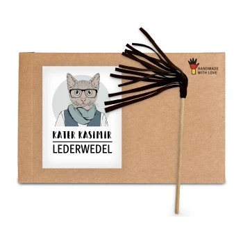 Ein Karton mit einer Cartoon-Katze mit Brille und Schal, beschriftet mit "Kater Kasimir Lederwedel", neben einem braunen Lederstabspielzeug.