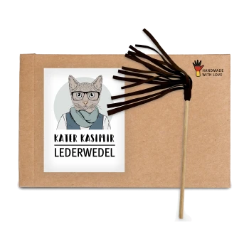 Ein Karton mit einer Cartoon-Katze mit Brille und Schal, beschriftet mit "Kater Kasimir Lederwedel", neben einem braunen Lederstabspielzeug.