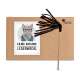 Ein Karton mit einer Cartoon-Katze mit Brille und Schal, beschriftet mit "Kater Kasimir Lederwedel", neben einem braunen Lederstabspielzeug.