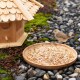 Ein kleiner brauner Vogel steht neben einem hölzernen Vogelhaus, das mit Samen gefüllt ist, auf einem Bett aus glatten Kieseln und grünem Moos.