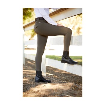 Eine Person in eng anliegenden olivfarbenen Reithosen und schwarzen Stiefeletten lehnt sich in einer sonnigen Außenumgebung an einen weißen Zaun.