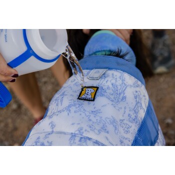 Eine Person gießt Wasser aus einer Flasche auf einen Hund, der eine blau-weiße Ruffwear-Kühlweste trägt, die für Outdoor-Aktivitäten konzipiert ist.