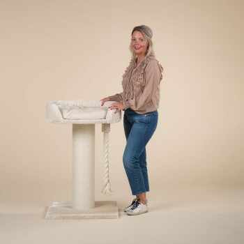 Eine Frau steht neben einem plüschigen Katzenbaum mit einem weichen Bett oben und einem Kratzbaum, vor einem neutralen Hintergrund.