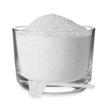 Eine klare Glasschüssel, gefüllt mit weißem Pulver, begleitet von einem kleinen weißen Messlöffel, der am Rand der Schüssel lehnt.