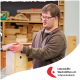 Eine Person in einem braunen Pullover montiert ein Holzvogelhaus in einer Werkstatt, mit dem Logo der Lebenshilfe Werkstätten e.V. in der Ecke.