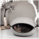 Ein graues Filz-Katzenbett mit einer getigerten Katze, die herausblickt, vor einem neutralen Hintergrund mit einer dekorativen Pflanze.