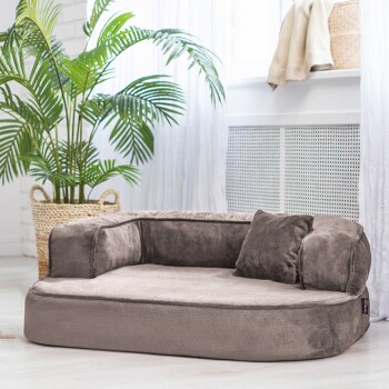 Ein plüschiges, graues Haustierbett mit niedrigem Profil und zwei seitlichen Kissen, platziert in einem hellen Raum mit einer Topfpflanze im Hintergrund.