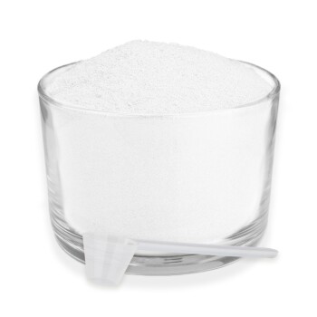 Ein klares Glasgefäß, gefüllt mit weißem Pulver, begleitet von einem kleinen weißen Messlöffel, der daneben ruht.