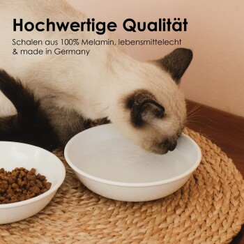 Eine Siamkatze trinkt aus einer weißen Melamin-Schüssel neben Trockenfutter auf einem Platzdeckchen. Text: "Hochwertig, lebensmittelecht, hergestellt in Deutschland."