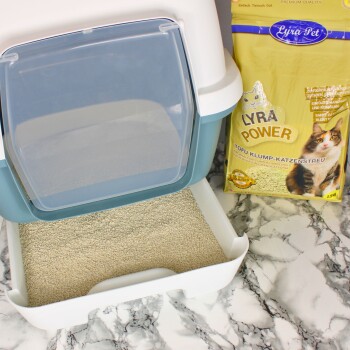Eine Katzenstreu-Box mit einer klaren Klapptür, gefüllt mit hellfarbiger Streu, neben einem Sack Lyra Power Tofu-Klumpenstreu.