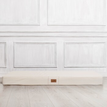 Ein cremefarbenes Haustierbett mit einem Lederetikett steht vor einer weißen Paneelwand und zeigt ein minimalistisches Design.