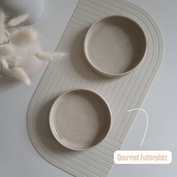 Zwei beige Keramik-Hundenäpfe auf einer hellen Matte, beschriftet mit "Gourmet Futterplatz," mit einer dekorativen Vase mit Trockenblumen in der Nähe.