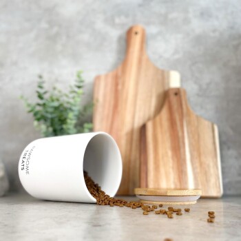 Ein weißer Behälter mit der Aufschrift "PAWSOME TREATS", der braune Haustierleckereien auf eine helle Arbeitsplatte auskippt, mit Holzschneidebrettern und Grünpflanzen im Hintergrund.