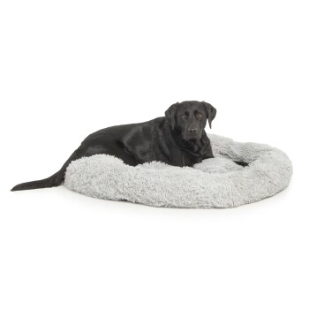 Ein schwarzer Labrador Retriever liegt auf einem flauschigen grauen Hundebett und schaut direkt in die Kamera.