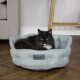 Eine schwarz-weiße Katze liegt in einem weichen, hellblauen Scruffs-Haustierbett, das auf einem Holzboden mit einem grauen Sofa im Hintergrund steht.
