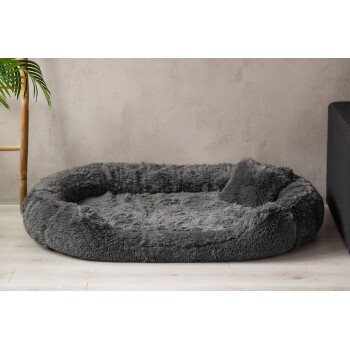 Ein gemütliches, rundes graues Haustierbett mit flauschiger Textur, auf einem hellen Holzboden neben einer Bambuspflanze und einem dunklen Sofa platziert.