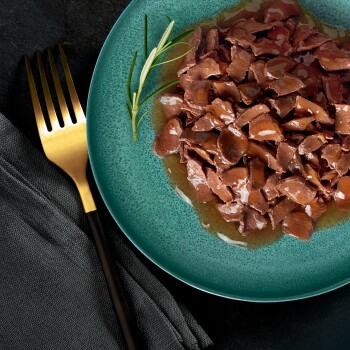 Zbliżenie na turkusowy talerz wypełniony rozdrobnioną karmą dla zwierząt, udekorowany gałązką rozmarynu, obok złotego widelca i czarnej serwetki.