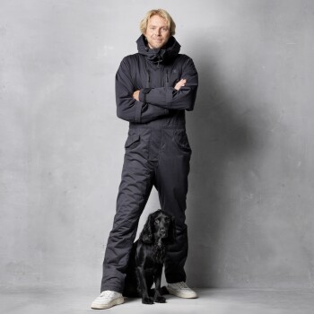 Eine Person in einem schwarzen Winteroverall steht selbstbewusst mit verschränkten Armen neben einem schwarzen Hund, vor einem grauen Hintergrund.