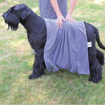 Eine Person legt ein graues Handtuch über einen großen schwarzen Hund, der auf Gras steht, und zeigt ein verwendetes Haustierpflegeprodukt.