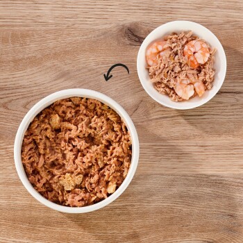 Eine große weiße Schüssel, gefüllt mit zerkleinertem Tierfutter, neben einer kleinen Schüssel mit zerkleinertem Futter mit Garnelenteilen, auf einer Holzoberfläche.