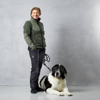 Eine Person in einer grünen Jacke und schwarzen Hosen steht neben einem großen, flauschigen schwarz-weißen Hund, beide vor einem grauen Hintergrund.