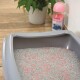Een grijze kattenbak gevuld met grijze en roze kattenbakvulling, met een groene plant op de achtergrond en een roze fles kattenbakvulling toevoeging in de buurt.