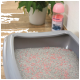 Une litière pour chat grise remplie de litière grise et rose, avec une plante verte en arrière-plan et une bouteille rose d'additif pour litière à proximité.