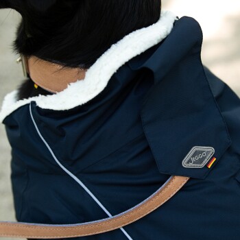 Eine Nahaufnahme des Halses eines schwarzen Hundes, der eine marineblaue Jacke mit einem weißen Fleecekragen trägt, mit einem Jeggo-Logo und einer braunen Lederleine.