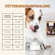 Ein Hund, der sich die Lippen leckt, neben einer Metallfutterschüssel, mit einer Verpackung von "WILDFANG Ente" Hundefutter und einer Fütterungsempfehlungstabelle auf Deutsch.