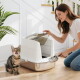 Frau öffnet weiße geschlossene Katzentoilette und schöpft Streu, während eine getigerte Katze daneben sitzt und sich die Lippen leckt.
