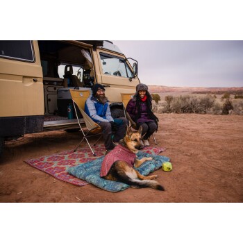 Ein Deutscher Schäferhund, der einen roten Mantel trägt, liegt auf einer blauen Decke vor einem Wohnmobil, während zwei Personen in einer Wüstenlandschaft in der Nähe sitzen.