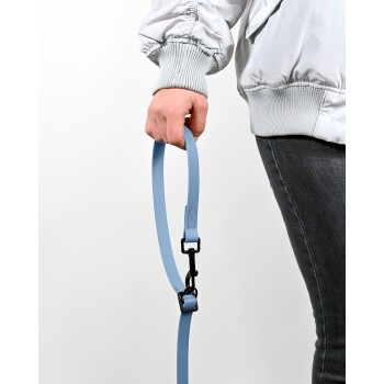 Eine Person hält eine hellblaue Hundeleine mit einem schwarzen Clip, trägt eine weiße Jacke und graue Jeans, vor einem einfarbigen Hintergrund.