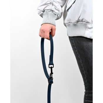Eine Person in einer hellgrauen Jacke hält eine dunkelblaue Haustierleine mit einem schwarzen Clip und steht vor einem weißen Hintergrund.