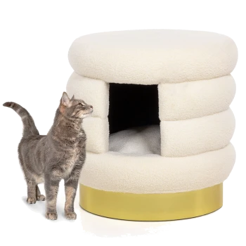 Eine graue Katze steht neben einem plüschigen, cremefarbenen Katzenbett mit runder Form und einem goldenen Sockel, das einen gemütlichen Eingang hat.