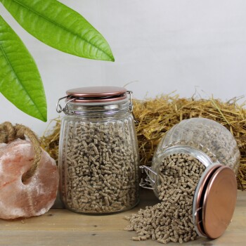 Zwei Glasgefäße mit Kupferdeckeln mit braunen Tierfutterpellets, eines aufrecht und eines verschüttet auf Holz; Stroh-Hintergrund.