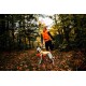 Eine Person in einer orange-schwarzen Jacke steht auf einem mit Blättern bedeckten Weg mit einem Hund, der eine passende orange-gelbe Weste trägt.