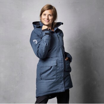 Eine Frau trägt eine blaue Outdoor-Jacke mit Kapuze, die zwei Fronttaschen und ein Logo am Ärmel hat, und posiert vor einem grauen Hintergrund.