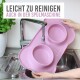 Eine Person hält eine rosa Silikon-Haustierfütterungsmatte mit zwei Schalen über einem Waschbecken mit fließendem Wasser. Text: "Einfach zu reinigen, spülmaschinenfest."