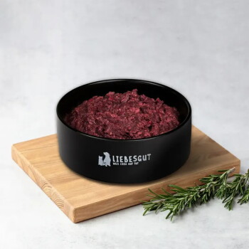 Schwarze Schale mit Aufschrift „LIEBESGUT (Weil Liebe Gut Tut)“, gefüllt mit rotem Rohfutter, auf einem Holzbrett, daneben ein Rosmarinzweig.