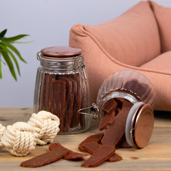 Ein Glasbehälter voller Hundeleckerlis, einige fallen auf einen Holztisch, neben einem geknoteten Seilspielzeug und einer rosa Haustiercouch im Hintergrund.