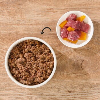 Eine weiße Schüssel mit feuchtem Haustierfutter steht auf einer Holzoberfläche, neben einem kleinen weißen Teller mit rohen Fleischstücken und gelben Paprikastücken.