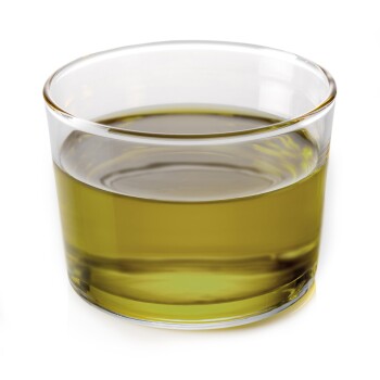 Ein klares Glas, gefüllt mit einem grünlichen Öl, das Licht reflektiert, auf einem weißen Hintergrund.