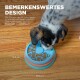 Ein schwarz-weißer Hund frisst Trockenfutter aus einem blauen Puzzle-Futterspender auf einem Holzboden. Deutscher Text lautet "BEMERKENSWERTES DESIGN."