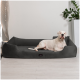 Ein weißer Hund, der auf einem plüschigen grauen Hundebett in einem modernen Innenraum entspannt, mit einer dekorativen Pflanze und Lampe im Hintergrund.