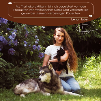 Eine Frau mit langen Haaren hält eine schwarze Katze neben einem großen Hund auf Gras, umgeben von lila Blumen. Der Text oben bewirbt Wolfsbacher Natur.