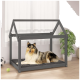 Ein Collie, der auf einem grauen Holz-Hundebett mit einem hausförmigen Rahmen entspannt, in einem hellen Raum mit einer Pflanze und Spielzeug in der Nähe.