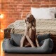Ein grauer Weimaraner-Hund sitzt aufrecht in einem plüschigen grauen Hundebett und schaut aufmerksam zur Seite, mit einem gemütlichen Bett und einer Ziegelwand im Hintergrund.