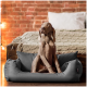 Ein grauer Weimaraner-Hund sitzt aufrecht in einem plüschigen grauen Hundebett und schaut aufmerksam zur Seite, mit einem gemütlichen Bett und einer Ziegelwand im Hintergrund.